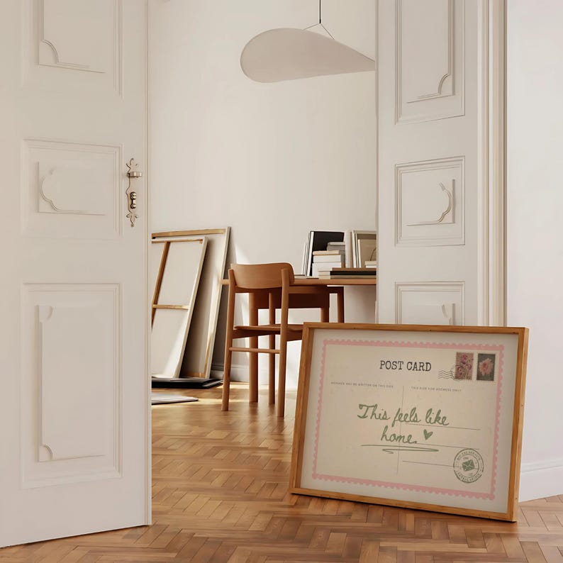 May include: A framed postcard with the text "This feels like home" in a wooden frame. The postcard has a pink border and features floral stamps. The scene is set in a room with a wooden floor and white doors.