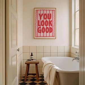 May include: A bathroom scene featuring a framed print with the words "YOU LOOK GOOD" in red against a pink striped background. A wooden stool holds a bottle and brush, and a white towel rests on the edge of a white bathtub. The floor has a black and white checkered pattern.