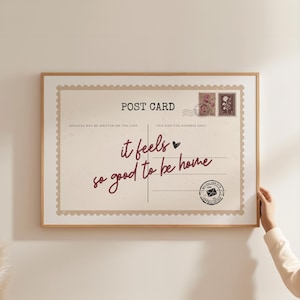 May include: A framed postcard print with a beige background and a wooden frame. The text "it feels so good to be home" is written in burgundy script. The print has a vintage aesthetic with postage stamp details.