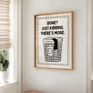 May include: A framed print with the text "DONE? JUST KIDDING. THERE'S MORE." above a black and white illustration of a laundry basket with a shirt and a dark cloth. The frame is a light brown color.