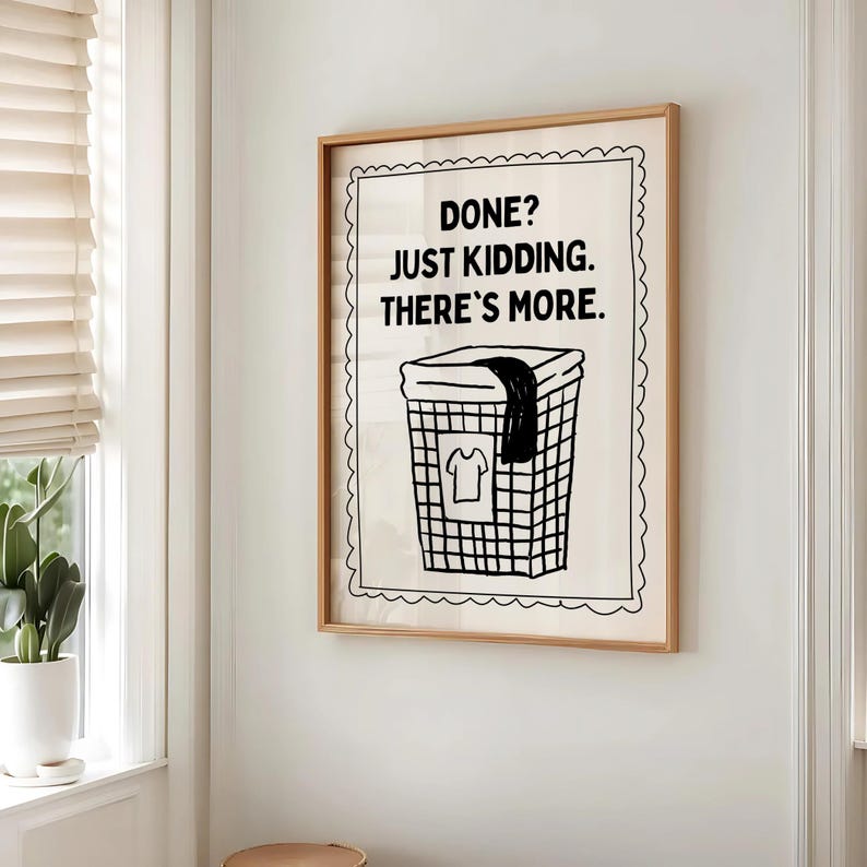 May include: A framed print with the text "DONE? JUST KIDDING. THERE'S MORE." above a black and white illustration of a laundry basket with a shirt and a dark cloth. The frame is a light brown color.