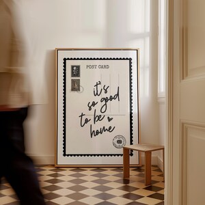 May include: A framed art print resembling a postcard, with the text "it's so good to be home" in cursive. The print has a black border and a gold frame. A small wooden stool sits nearby on a checkered floor.