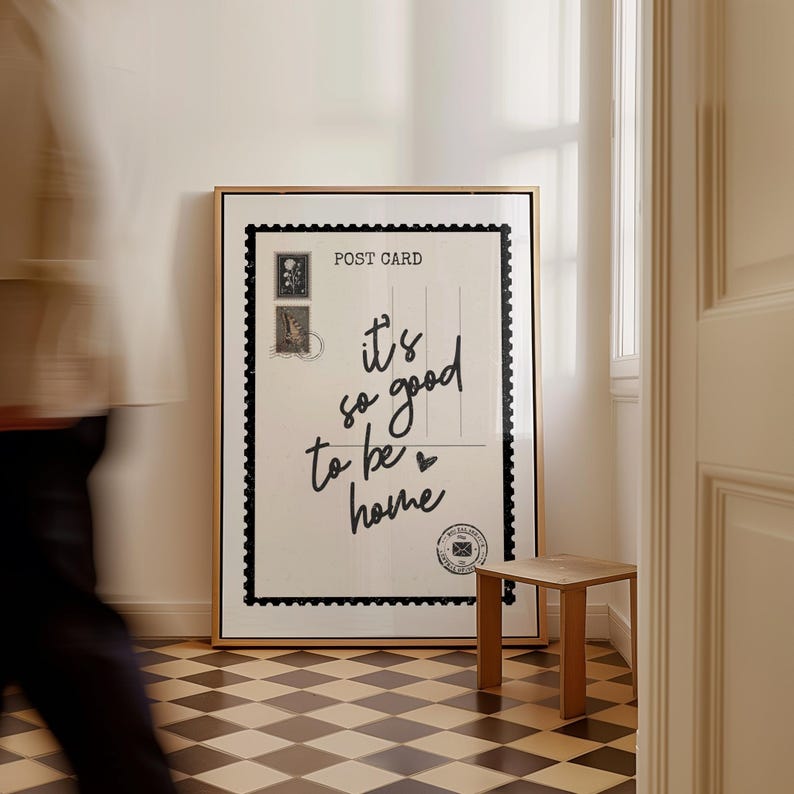 May include: A framed art print resembling a postcard, with the text "it's so good to be home" in cursive. The print has a black border and a gold frame. A small wooden stool sits nearby on a checkered floor.