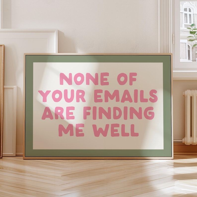 May include: A framed art print with the text "NONE OF YOUR EMAILS ARE FINDING ME WELL" in pink letters. The print has a light green border and a beige background. The frame is a light brown color.