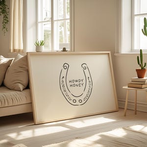 May include: A framed art print featuring a black horseshoe design with the words "HOWDY HONEY" in the center. The artwork is displayed on a cream-colored background, set in a light wooden frame. The print is leaning against a wall.