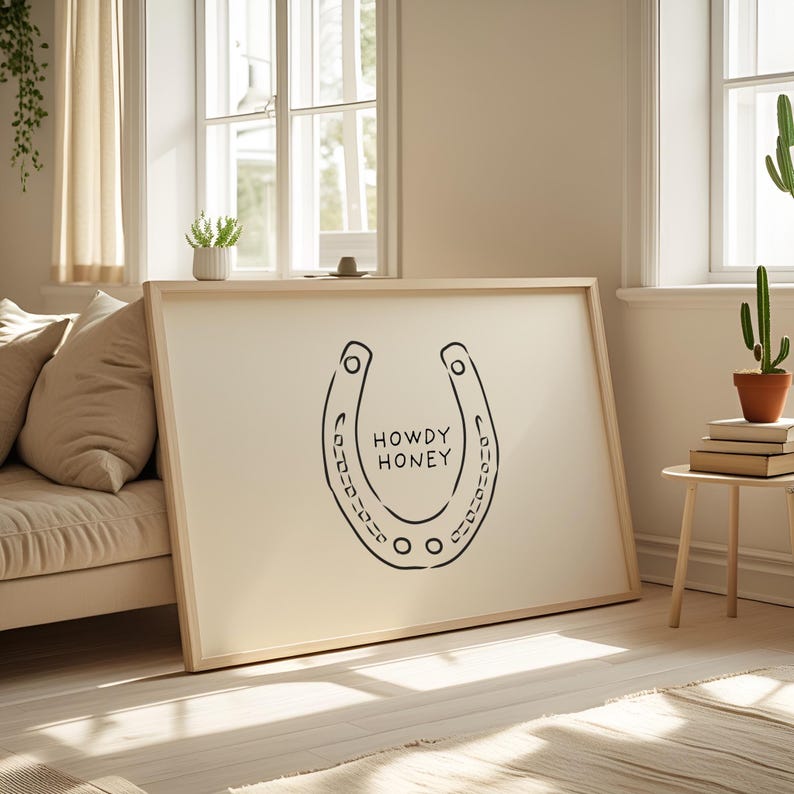May include: A framed art print featuring a black horseshoe design with the words "HOWDY HONEY" in the center. The artwork is displayed on a cream-colored background, set in a light wooden frame. The print is leaning against a wall.