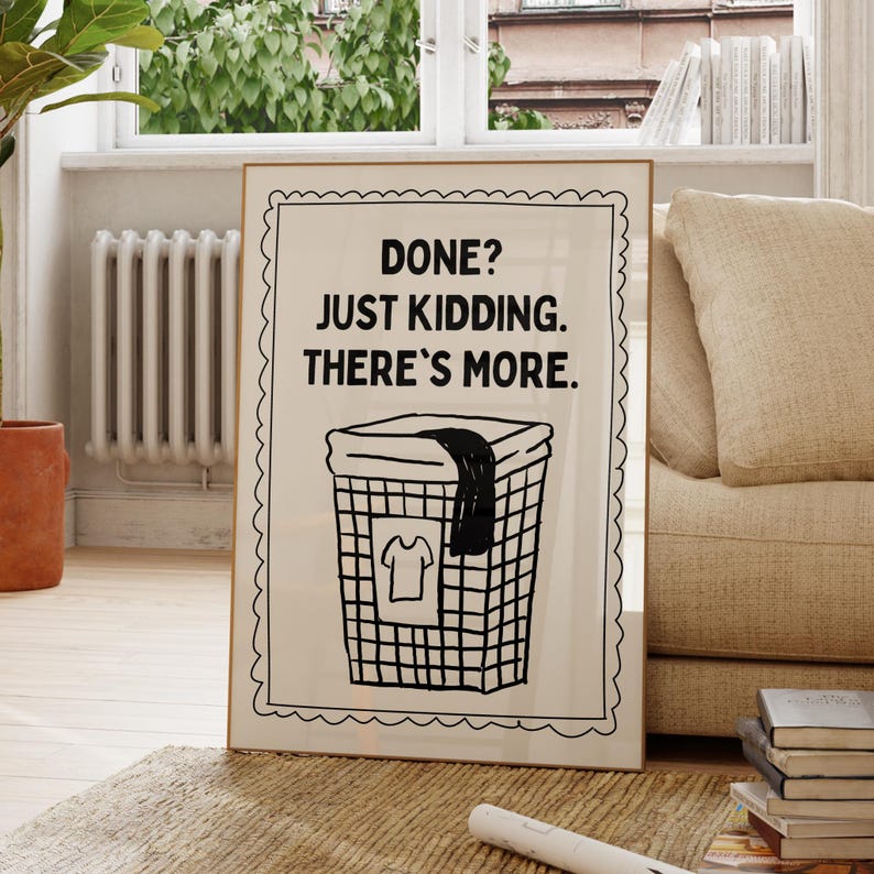 May include: A framed print with the text "DONE? JUST KIDDING. THERE'S MORE." features a black and white illustration of a laundry basket with a shirt inside. The frame has a decorative border and is displayed in a living room setting.