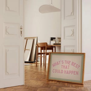 May include: A framed print with the text "WHAT'S THE BEST THAT COULD HAPPEN" in pink lettering. The print has a light green border and a wooden frame. The image is in a room with a wooden floor and white walls.