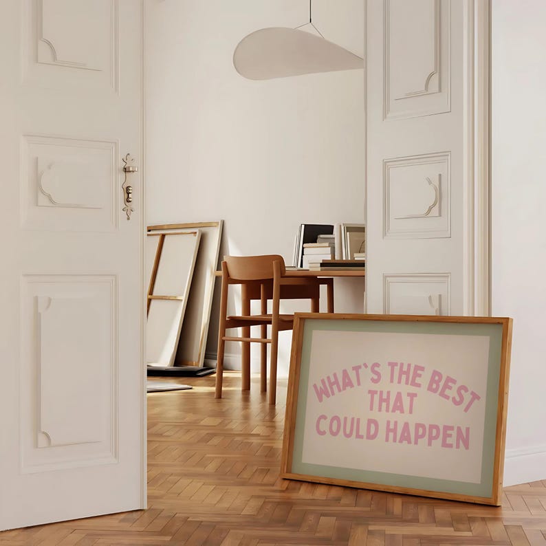 May include: A framed print with the text "WHAT'S THE BEST THAT COULD HAPPEN" in pink lettering. The print has a light green border and a wooden frame. The image is in a room with a wooden floor and white walls.