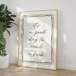 Puede incluir: Un estampado enmarcado con el texto "It's a good day to read a book" en escritura negra. El estampado tiene un fondo color crema con un borde negro decorativo y está enmarcado en oro. Una gran planta en maceta es visible en el fondo.