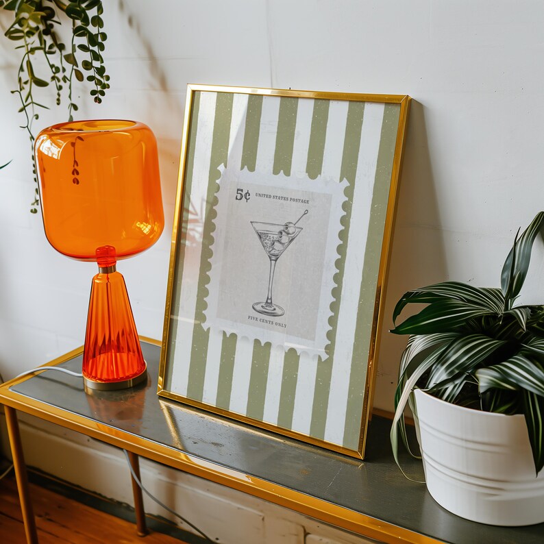 Op de afbeelding: Een oranje glazen lamp met een geribbelde voet en een rechthoekige kap staat op een goudomrande tafel. Daarnaast een ingelijste print met een gouden rand, met een martini-illustratie op een postzegelontwerp. De achtergrond heeft groene en witte strepen.