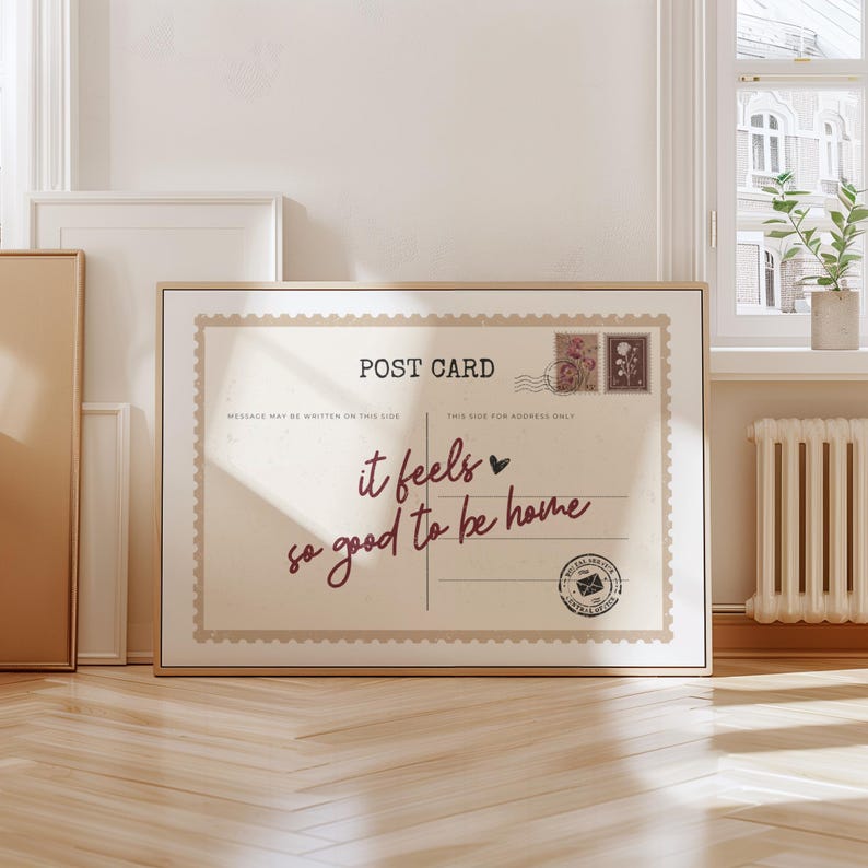 May include: A framed postcard print with a beige background and a vintage design. The text "POST CARD" is at the top, with the phrase "it feels so good to be home" in a handwritten font. Decorative stamps and a postal seal are also included.