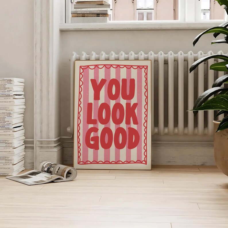 May include: A framed art print with the words "YOU LOOK GOOD" in red block letters on a pink and white striped background. The print has a scalloped border and is displayed on a light wood floor.