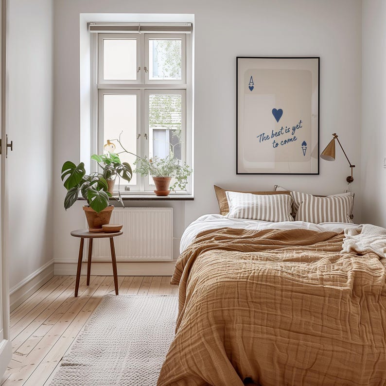 May include: A bedroom scene with a bed covered in a mustard-colored quilt and striped pillows. A framed print with the text "The best is yet to come" hangs above the bed. A small wooden side table holds a potted plant, and a window provides natural light.