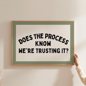 May include: A framed art print with the text "DOES THE PROCESS KNOW WE'RE TRUSTING IT?" in black lettering. The frame is a light wood color with a green border, and the background is off-white.