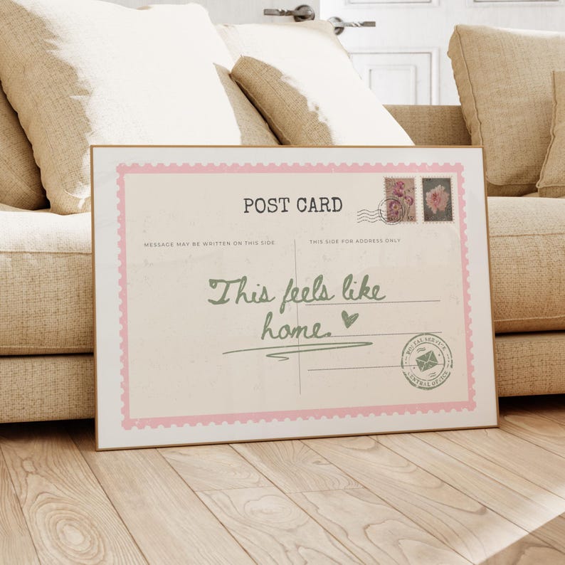 May include: A framed postcard with a gold border and a pink scalloped edge. The postcard reads "POST CARD" at the top, with the text "This feels like home" in cursive. Two floral stamps and a postal seal are also visible.