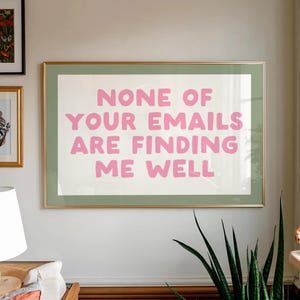 Op de afbeelding: Een ingelijste kunstdruk met de tekst "NONE OF YOUR EMAILS ARE FINDING ME WELL" in roze letters. De print heeft een gouden lijst en een groene passe-partout. Het kunstwerk wordt op een witte muur getoond.