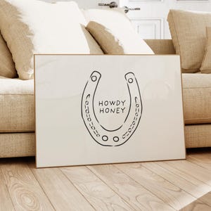 May include: A framed print featuring a black horseshoe design with the words "HOWDY HONEY" in the center. The artwork has a minimalist style and is set against a cream-colored background. The frame is a light gold color.