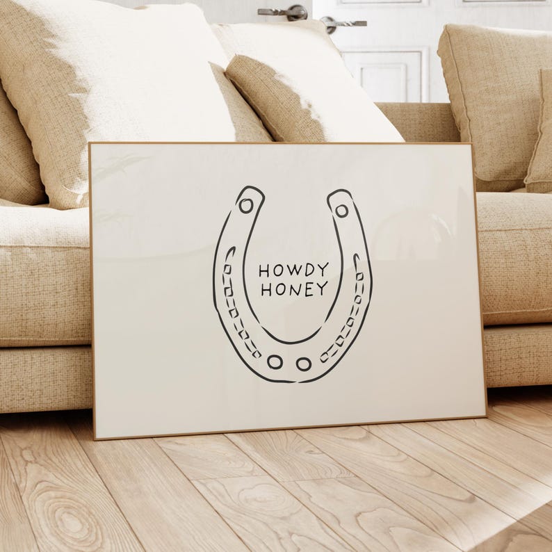 May include: A framed print featuring a black horseshoe design with the words "HOWDY HONEY" in the center. The artwork has a minimalist style and is set against a cream-colored background. The frame is a light gold color.