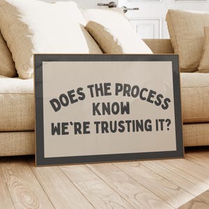 May include: Framed art print with the text "DOES THE PROCESS KNOW WE'RE TRUSTING IT?" in a black font. The print has a beige background and a black frame. The artwork is displayed on a wooden floor.