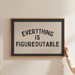 Pode incluir: Uma impressão emoldurada com o texto "EVERYTHING IS FIGUREOUTABLE" em preto sobre um fundo creme. A moldura é de madeira clara com uma borda preta. A impressão está sendo segurada por uma pessoa.