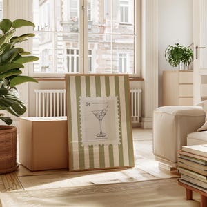 Op de afbeelding: Een ingelijste print met een cocktailillustratie op een postzegelontwerp. De lijst heeft een lichtbruine rand en een groen-witte gestreepte achtergrond. De print leunt tegen een bruine kubusvormige doos op een houten vloer.