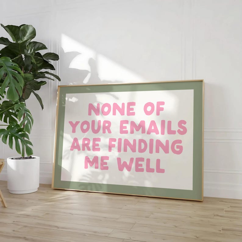 May include: A framed art print with the text "NONE OF YOUR EMAILS ARE FINDING ME WELL" in pink letters. The print has a white background and a green border, set in a gold frame. A potted plant is visible in the background.