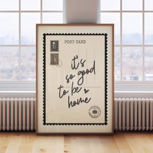 May include: A framed postcard print with the text "it's so good to be home" in a handwritten style. The print has a vintage aesthetic with postage stamps and a black border, set against a neutral background.