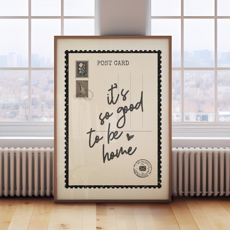 May include: A framed postcard print with the text "it's so good to be home" in a handwritten style. The print has a vintage aesthetic with postage stamps and a black border, set against a neutral background.