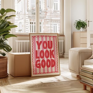 May include: A framed art print with the words "YOU LOOK GOOD" in bold, pink letters on a striped background. The print has a scalloped border and is displayed in a bright, airy room with a large window and a potted plant.