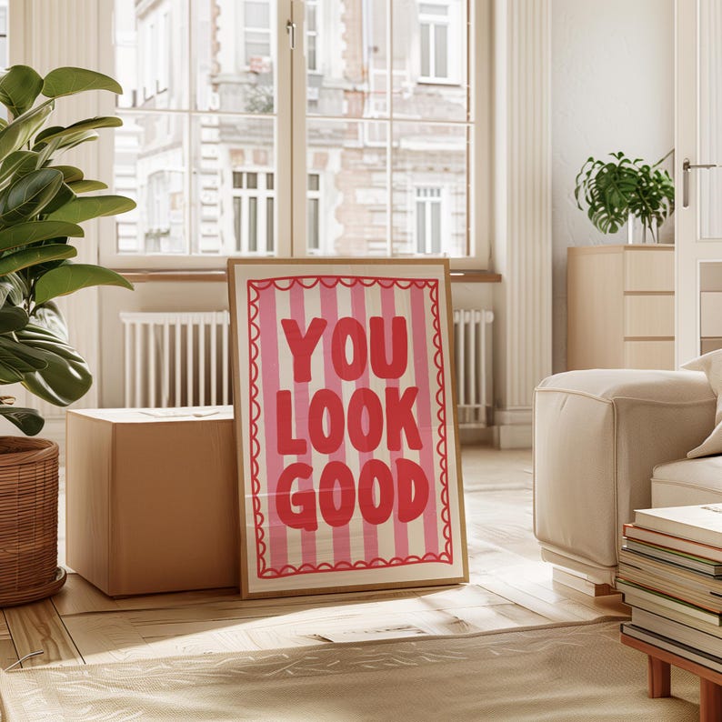 May include: A framed art print with the words "YOU LOOK GOOD" in bold, pink letters on a striped background. The print has a scalloped border and is displayed in a bright, airy room with a large window and a potted plant.