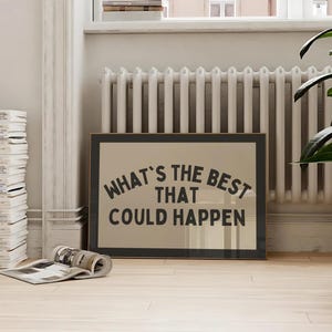 May include: A framed print with the text "WHAT'S THE BEST THAT COULD HAPPEN" in a black, bold font. The print is displayed on a light-colored wooden floor, with a stack of magazines and a potted plant in the background.
