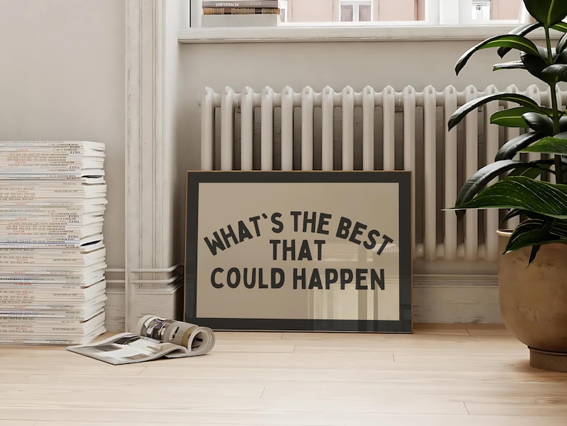 May include: A framed print with the text "WHAT'S THE BEST THAT COULD HAPPEN" in a black, bold font. The print is displayed on a light-colored wooden floor, with a stack of magazines and a potted plant in the background.