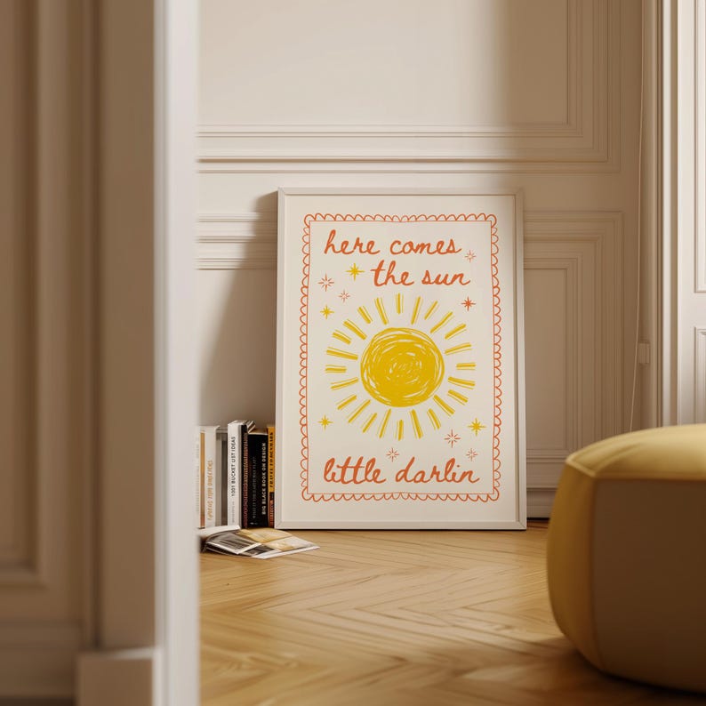 May include: A framed art print with the words "here comes the sun" and "little darlin" in orange script. The artwork features a yellow sun illustration with rays and star accents, set against a cream background.
