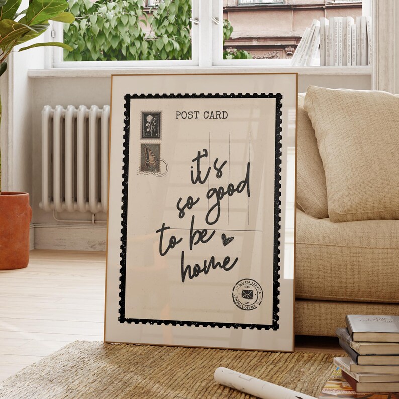 May include: A framed postcard print with the text "it's so good to be home" in black script. The print has a postage stamp border and two stamp illustrations. The frame is gold, and the print is displayed indoors.
