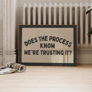May include: A framed print featuring the text "DOES THE PROCESS KNOW WE'RE TRUSTING IT?" in a dark, bold font. The print has a neutral background and a black frame. A rolled magazine lies in front of the print.