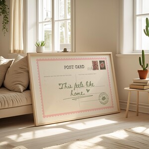May include: A framed postcard print with the text "This feels like home." The postcard has a pink scalloped border and decorative stamps. The frame is light-colored wood, and the print is leaning against a sofa.