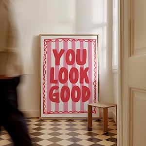 May include: A framed art print with the words "YOU LOOK GOOD" in red, set against a pink and white striped background. The artwork is in a gold frame and hangs on a white wall. A small wooden stool sits nearby.