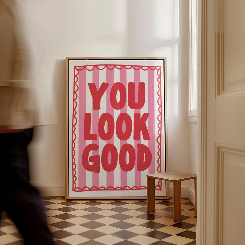 May include: A framed art print with the words "YOU LOOK GOOD" in red, set against a pink and white striped background. The artwork is in a gold frame and hangs on a white wall. A small wooden stool sits nearby.