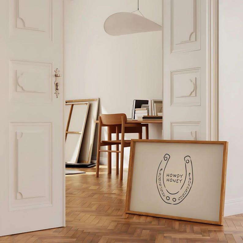 May include: A framed artwork featuring a black horseshoe design with the words "HOWDY HONEY" in a handwritten font. The artwork has a light beige background and a natural wood frame. The artwork is leaning against a wall in a room with hardwood floors.