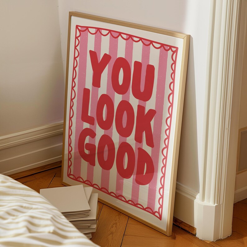 May include: A framed art print with the words "YOU LOOK GOOD" in large red letters on a pink and white striped background. The print has a scalloped pink border and a gold frame. The artwork is leaning against a white wall.