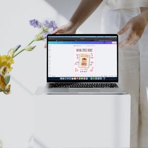 Pode incluir: Uma tela de laptop exibindo uma maquete de capa de livro com o texto "BOOK TITLE HERE" e um fundo rosa. A capa apresenta uma ilustra&ccedil;&atilde;o de desenho animado de uma pessoa com um livro e o texto "Little Words, Big Magic".