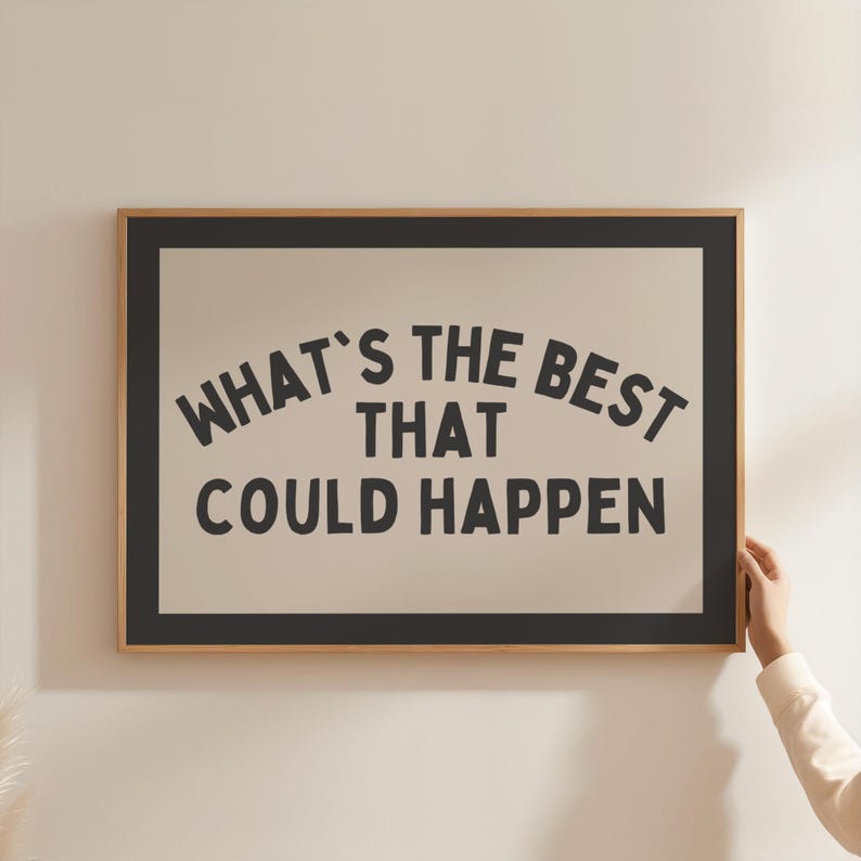 May include: A framed art print with the text "WHAT'S THE BEST THAT COULD HAPPEN" in black, centered on a beige background. The frame is a light wood color with a black border. The image is being held by a person.