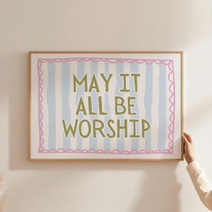 May include: A framed art print with the words "MAY IT ALL BE WORSHIP" in green lettering. The background has blue and white vertical stripes, and a pink scalloped border. The frame is made of light brown wood.