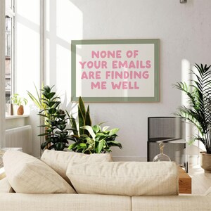 May include: A framed print with the text "NONE OF YOUR EMAILS ARE FINDING ME WELL" in pink letters. The print is in a green frame and hangs on a white wall. The room has a beige sofa, plants, and a window.