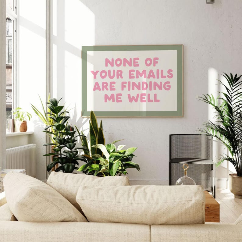 May include: A framed print with the text "NONE OF YOUR EMAILS ARE FINDING ME WELL" in pink letters. The print is in a green frame and hangs on a white wall. The room has a beige sofa, plants, and a window.