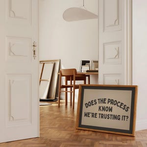 May include: A framed sign with the text "DOES THE PROCESS KNOW WE'RE TRUSTING IT?" in a room with a wooden floor. The sign is in a wooden frame with a black border. The room has white walls and a wooden desk and chair.