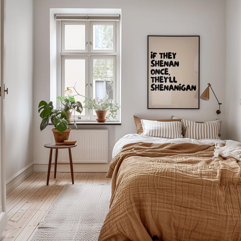 May include: A bedroom scene with a bed covered in a mustard-colored quilt and striped pillows. A framed poster with the text "IF THEY SHENAN ONCE, THEY'LL SHENANIGAN" hangs on the wall. A small table holds a potted plant, and a window provides natural light.