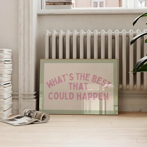May include: A framed print with the text "WHAT'S THE BEST THAT COULD HAPPEN" in pink lettering. The print is leaning against a wall near a radiator and a potted plant. A stack of magazines and a rolled-up magazine are in the foreground.