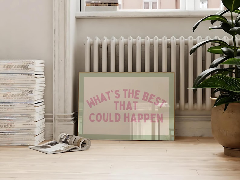 May include: A framed print with the text "WHAT'S THE BEST THAT COULD HAPPEN" in pink lettering. The print is leaning against a wall near a radiator and a potted plant. A stack of magazines and a rolled-up magazine are in the foreground.